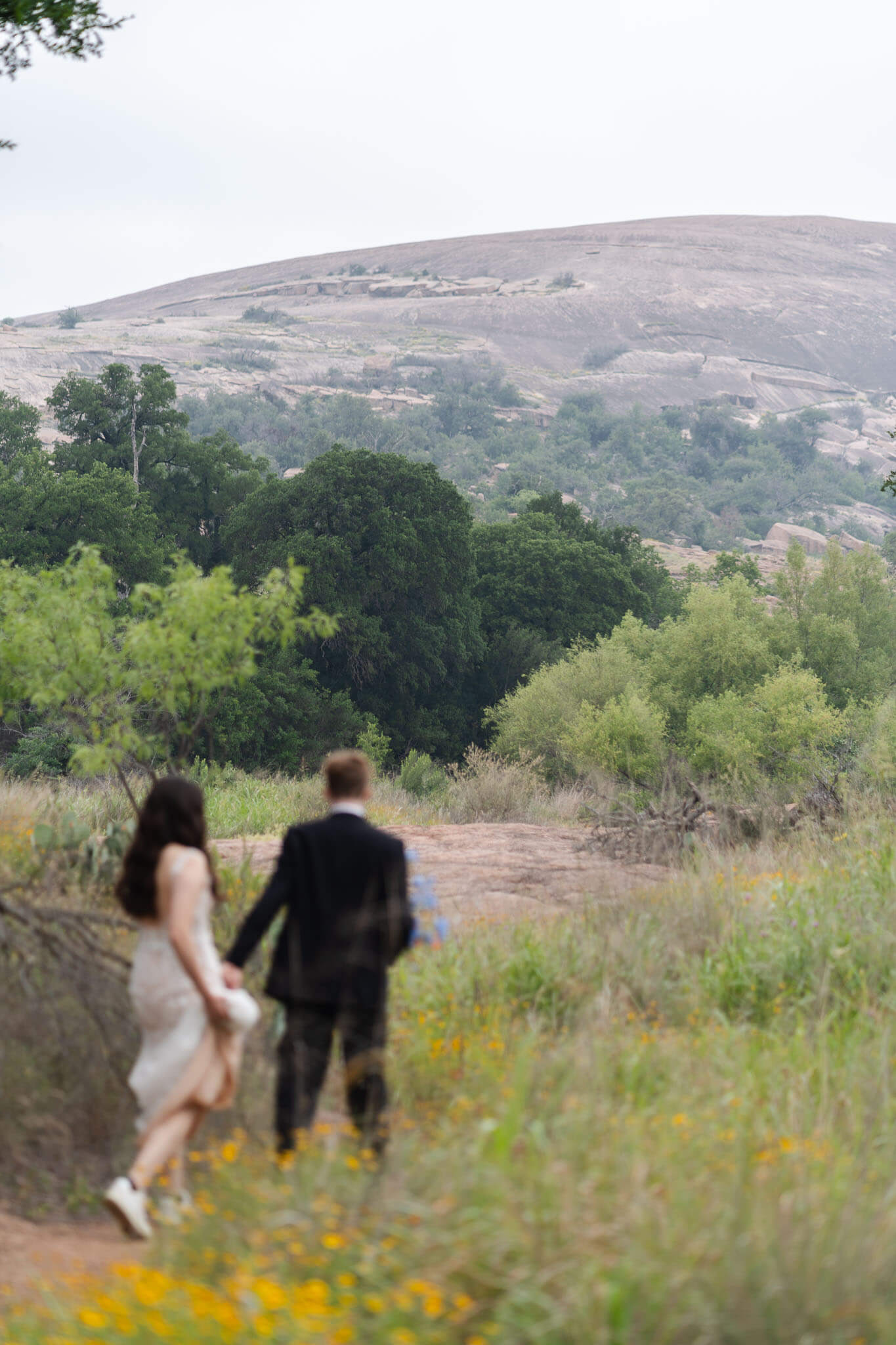 couple walks through a secluded trail lined with wildflowers with enchanted rock in front of them 
