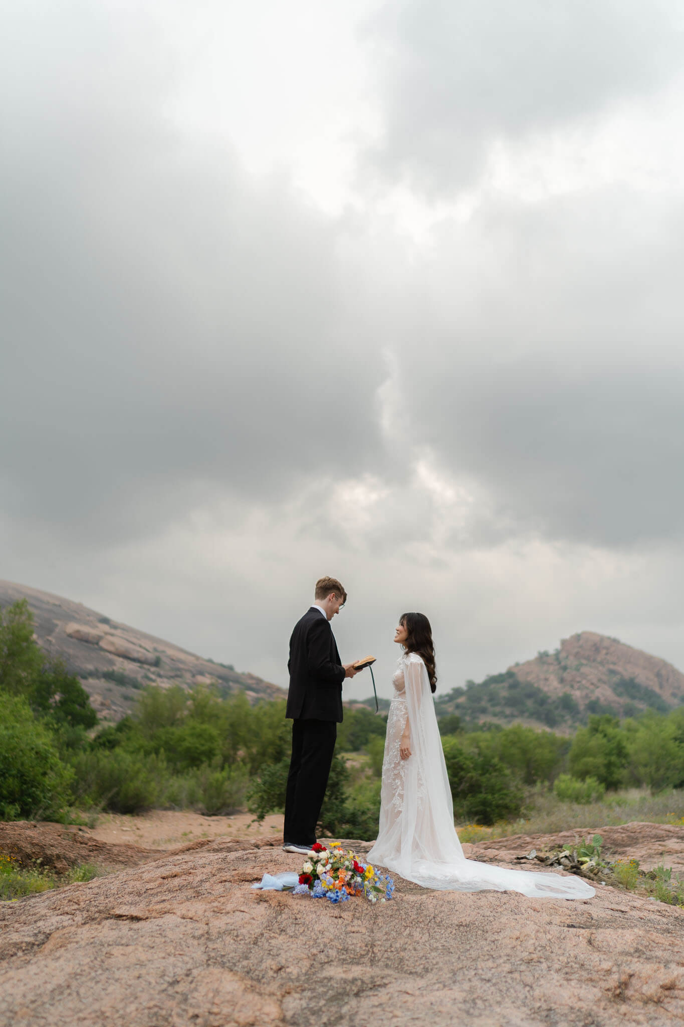 Bride and groom standing at the base of of Enchanted Rock at sunrise, holding hands, with pink granite dome behind them