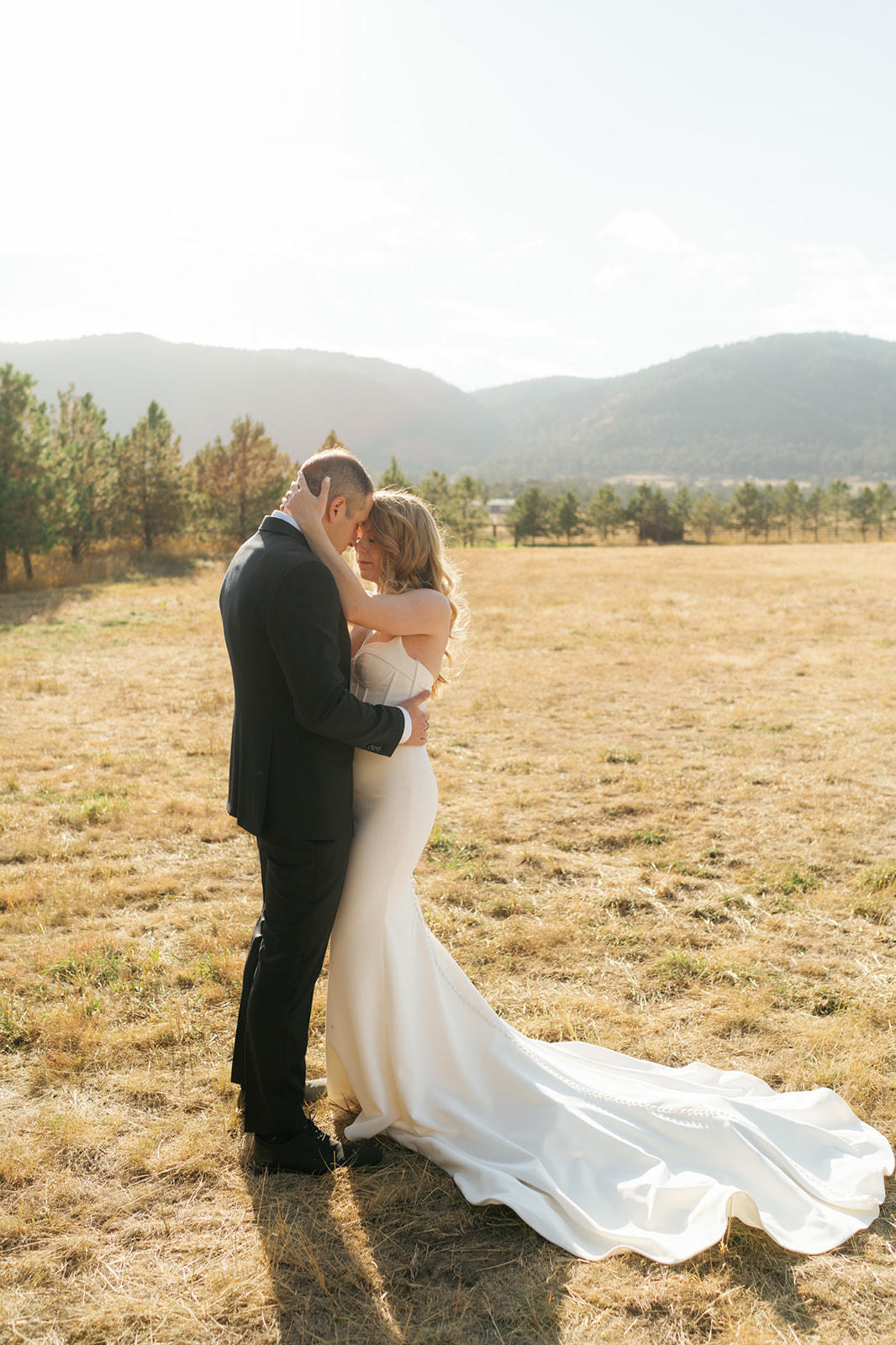 Newlyweds snuggle touching foreheads in an open pasture lined with trees and mountains