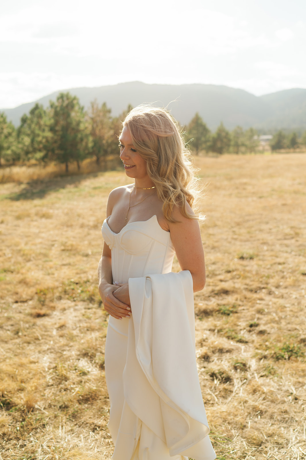 A bride holds her train over her arm while standing in a windy pasture with mountains behind her