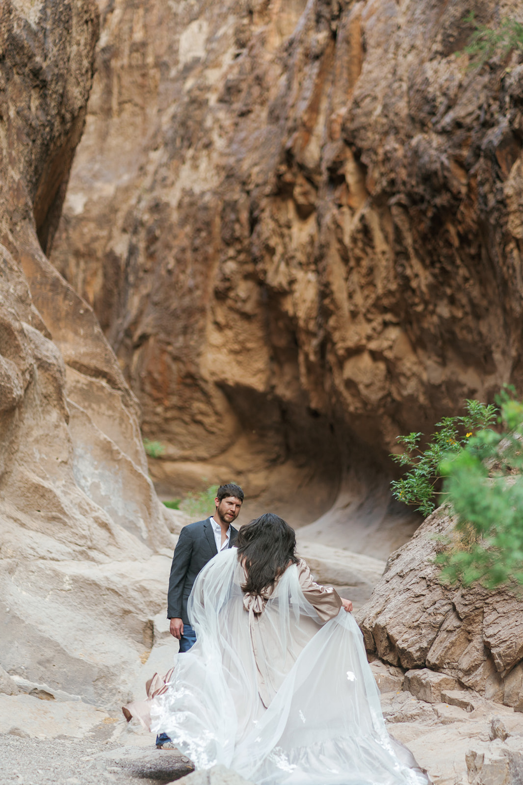A bride holds her long train while walking up to her groom in a tall canyon
