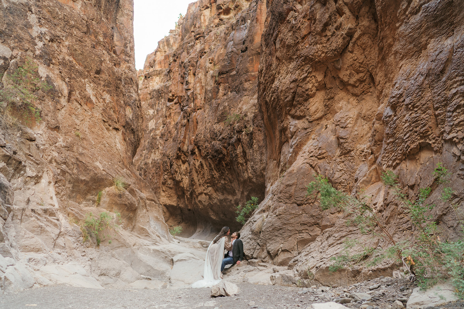 A bride leans in to kiss her sitting groom in a sandy canyon