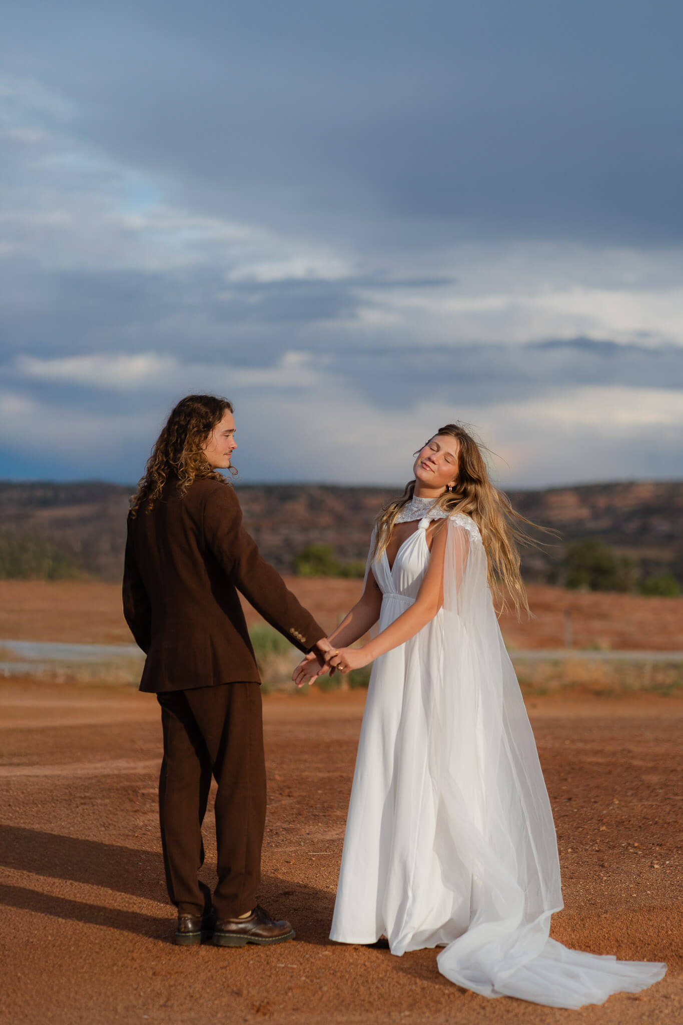 Emilie smiling toward the sun as Max watches, with a vibrant rainbow over Moab red rocks behind them during their Dead horse point wedding