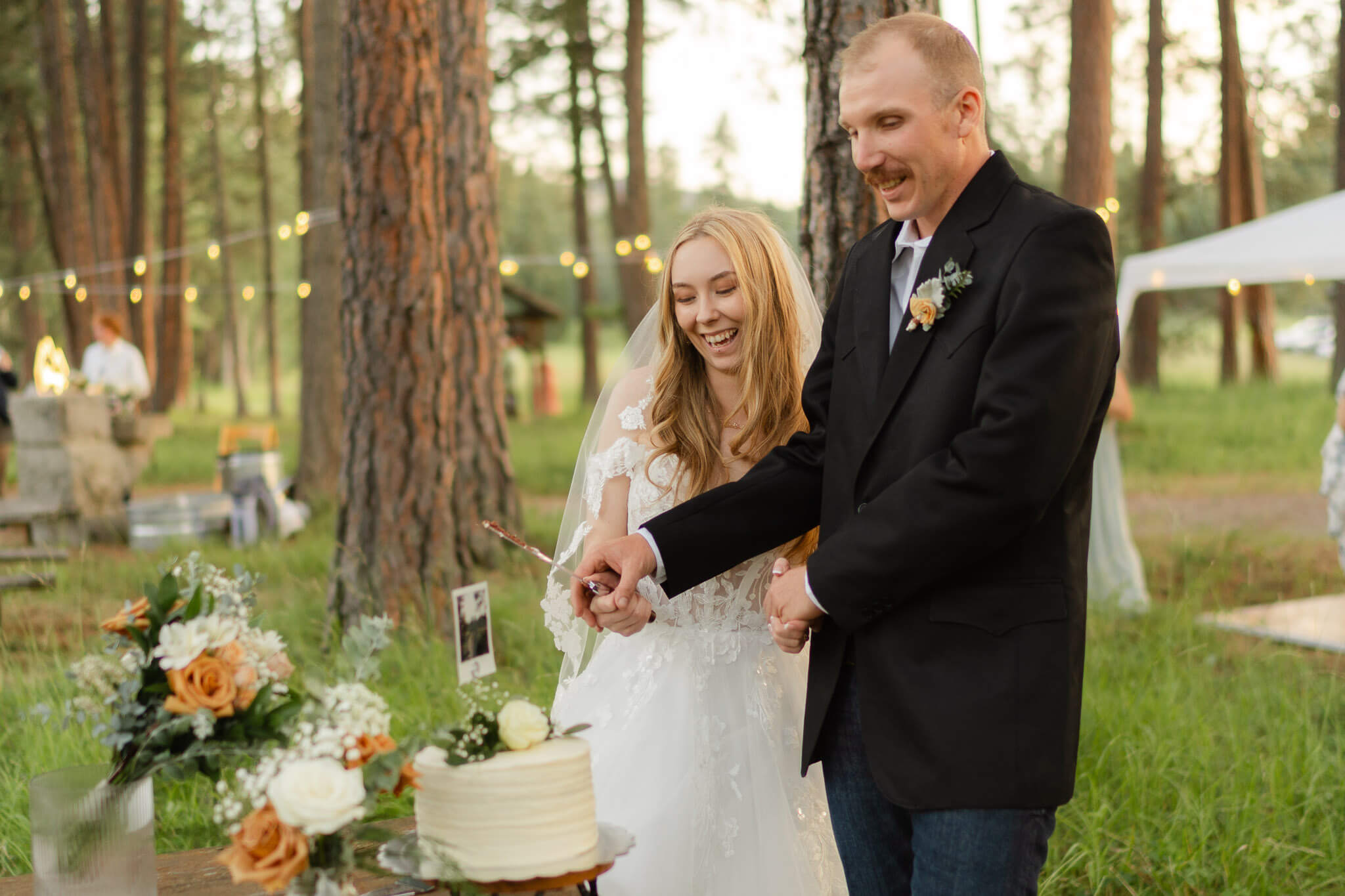 Bride and groom cut their wedding cake under forest canopy and string lights during their Bigfork wedding | Kalispell wedding photographer