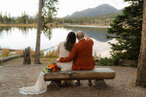 groom points off into the distance as couple sits together while the sun sets over a lake where they chose to elope in Colorado
